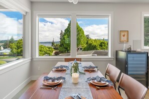 A dining room with a table set for four, large windows with a view of greenery and a body of water, and a small refrigerator on the right. - Modern Hilltop Home Lake Views ~ Decks & Patio! (Seattle)