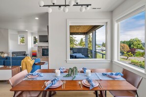 A dining area with a wooden table set for four, a large window with a view of the outdoors, and a modern living space in the background. - Modern Hilltop Home Lake Views ~ Decks & Patio! (Seattle)