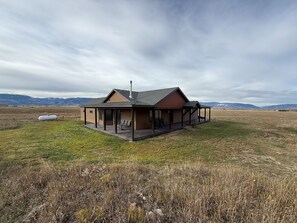 A single-story cabin with a deck, surrounded by a grassy field and mountains in the background. - Yellowstone Gateway Retreat: Scenic Paradise Valley Oasis on 20 Acres (Livingston)