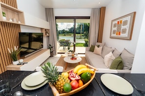 Ein modernes Wohnzimmer mit Fernseher, einem Essbereich mit Obstschale und Blick auf einen Garten durch große Fenster.
