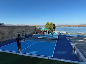 Una cancha de tenis con red, rodeada por una cerca de madera, y vista a un lago con montañas al fondo.