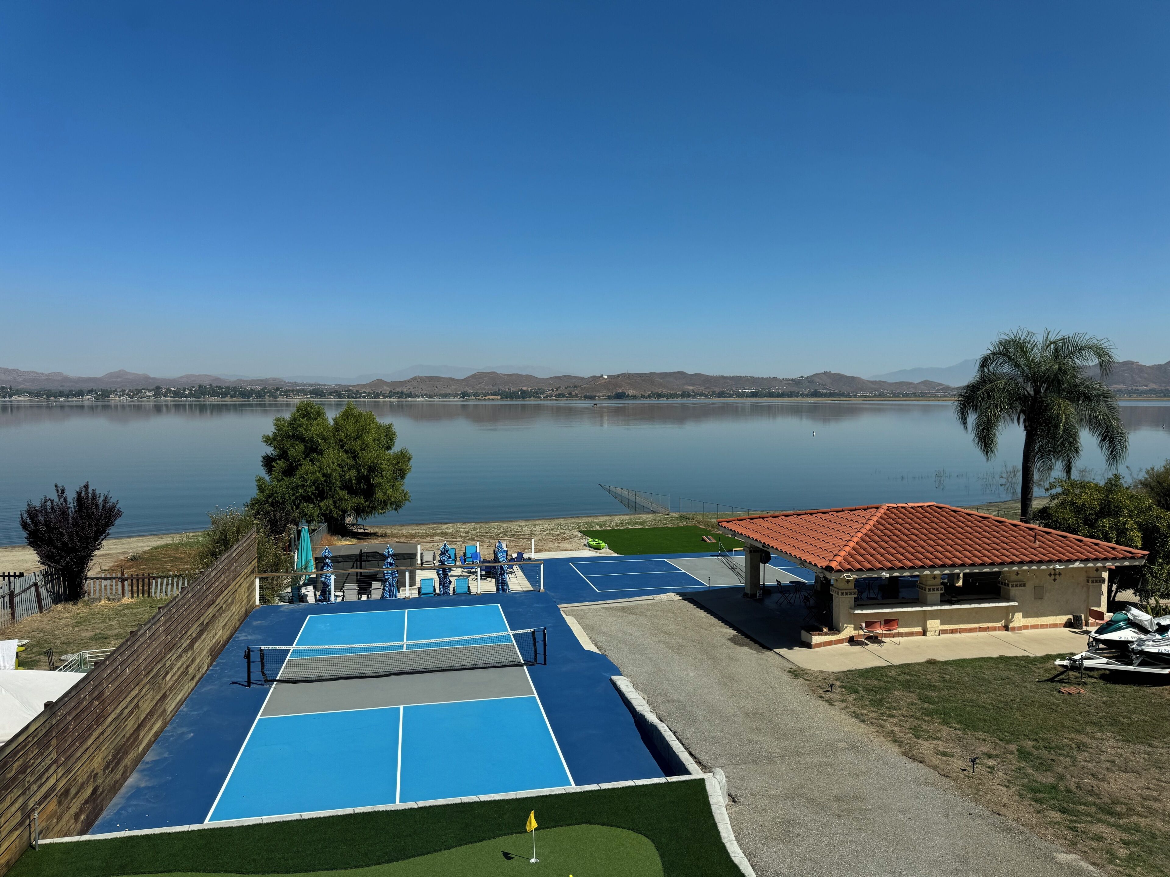 Ein Tennisplatz mit Netz neben einem Gebäude mit rotem Dach, mit Blick auf einen stillen See und Berge im Hintergrund.