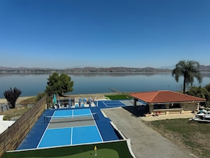 A tennis court with a net, adjacent to a building with a red roof, overlooking a calm lake with mountains in the distance.