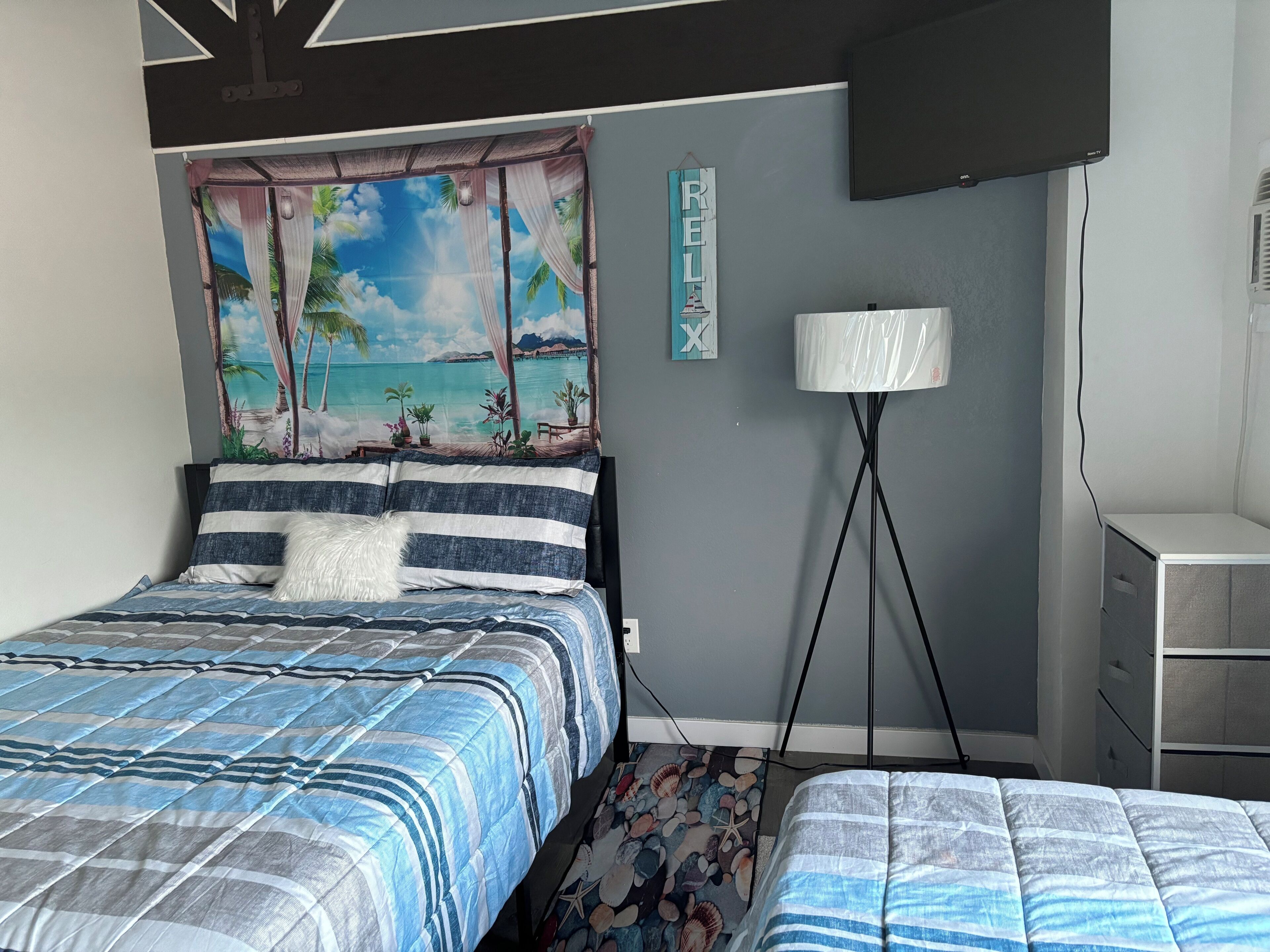 A bedroom with a bed, a nightstand, a lamp, and a wall with a tropical mural.
