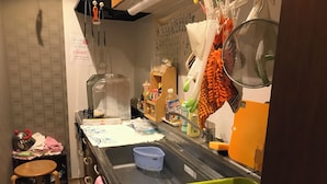 Una pequeña cocina con un lavamanos, una tetera, un reposa-teteras, un tendedero, una cesta verde y un taburete de madera.