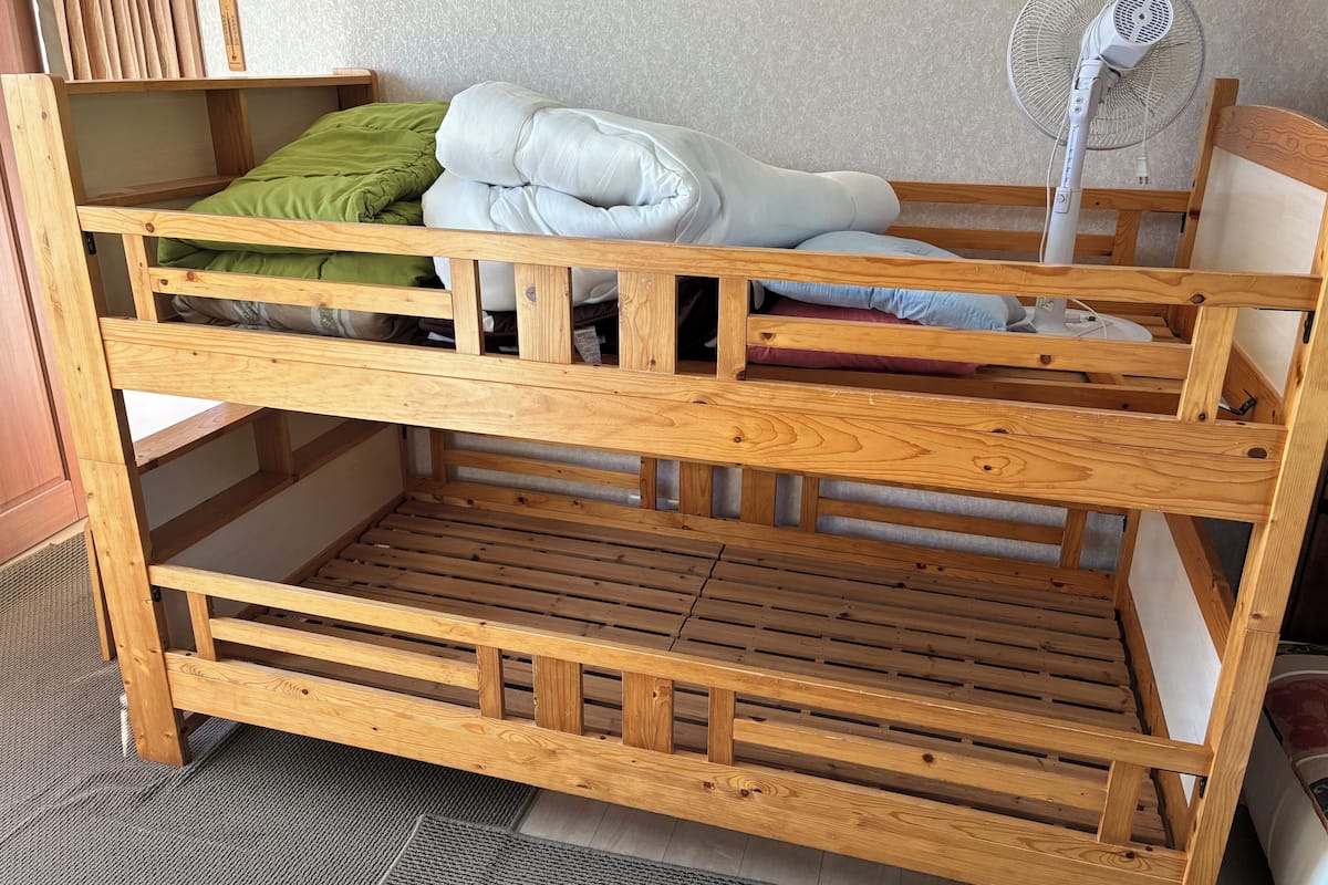 A wooden bunk bed with a fan and pillows, a wooden shelf, and a carpeted floor.