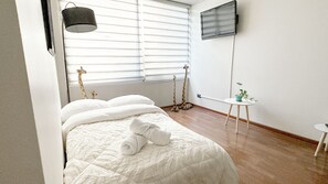 Ein Schlafzimmer mit einem Bett, einem kleinen Tisch, einer Topfpflanze und einem an der Wand befestigten Fernseher.