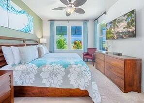 Habitación de hotel con cama, escritorio, silla, ventilador de techo y una ventana grande con vistas a la vegetación.