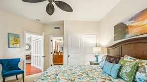 A bedroom with a bed, a ceiling fan, a nightstand, a lamp, a chair, and a door leading to another room. - Ocean View 6BR, 2 Condos for 16 in Koloa! (Koloa)