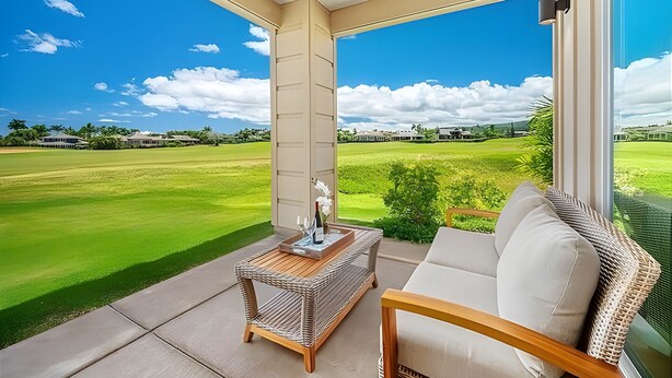 A balcony with a view of a golf course, a wicker table with a bottle and glasses, and a cushioned chair. - Ocean View 5BR, 2 Condos for 14 in Koloa! (Koloa)