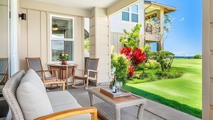 A covered outdoor seating area with wicker furniture, a table with a vase of flowers, and a view of a well-maintained lawn and house. - Ocean View 5BR, 2 Condos for 14 in Koloa! (Koloa)