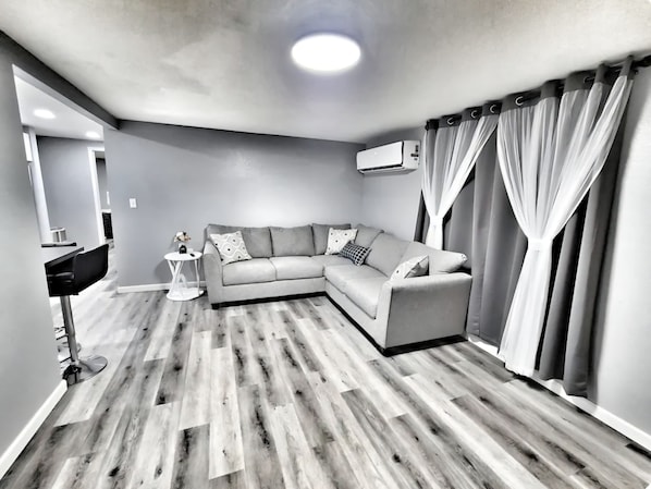 Un salon moderne avec un canapé gris, une petite table d’appoint et un coin repas à l’arrière-plan.