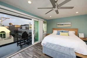 Ein Schlafzimmer mit Bett, Deckenventilator, Nachttisch, einer Bank und einer Schiebetür zu einem Außenbereich.