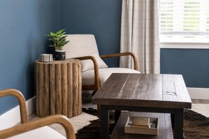 Ein Zimmer mit einem Holztisch, einem runden Beistelltisch, einem Sessel und einem Fenster mit Vorhängen.