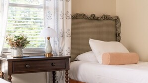 Un dormitorio con cama, mesita de noche con lámpara, un florero con flores y una ventana con cortinas.