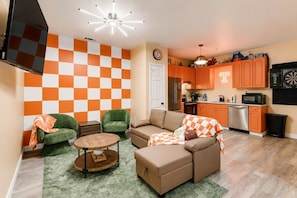 A compact living space with a checkered wall, a sofa, a small round table, and a kitchen area with orange cabinets and modern appliances. - Game Day Getaway | Vols-Themed Stay Near Vanderbilt (Nashville)