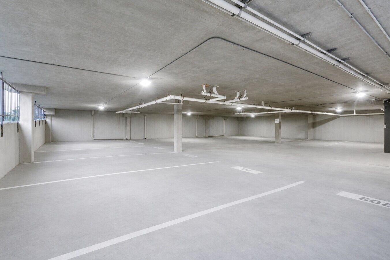 Eine leere Parkgarage mit Betonböden und -wänden, einer weißen Deckeninstallation und einigen sichtbaren Säulen.