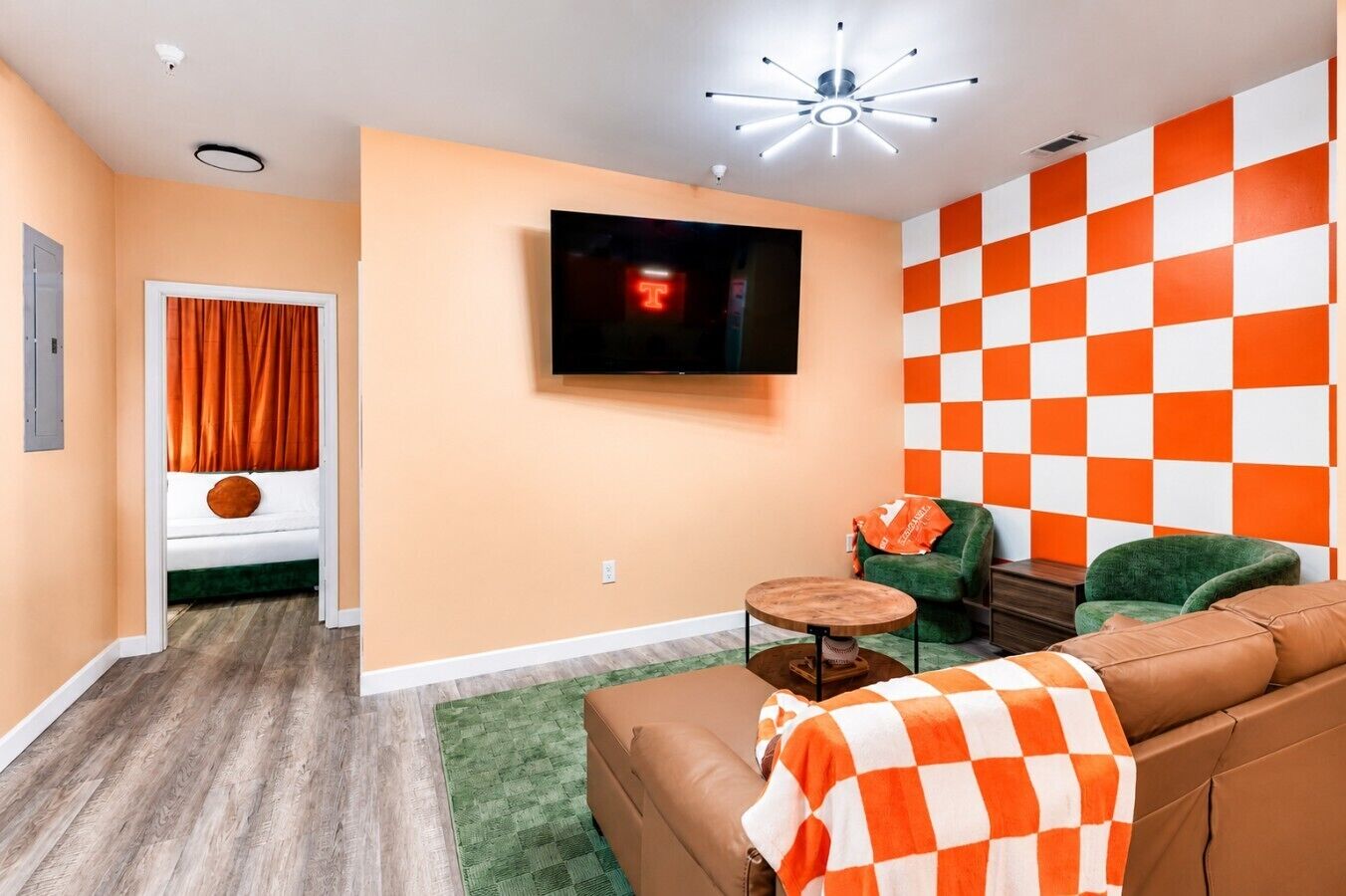 Una sala de estar con televisor, pared de cuadros naranjas y blancos, sillón verde, sofá marrón y una pequeña mesa redonda.