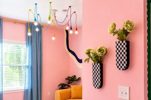 Ein Zimmer mit einer rosafarbenen Wand, einem Fenster mit blauen Vorhängen, einem gelben Sofa, einer karierten Vase mit Blumen und einer mehrfarbigen Leuchte.