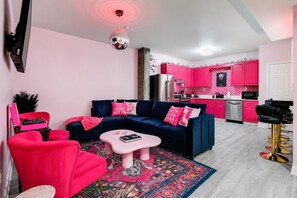 Ein Wohnzimmer mit blauem Sofa, rosa Sessel und Couchtisch. Eine Küche mit rosafarbenen Schränken und ein Essbereich mit einem schwarzen Barhocker sind ebenfalls zu sehen.