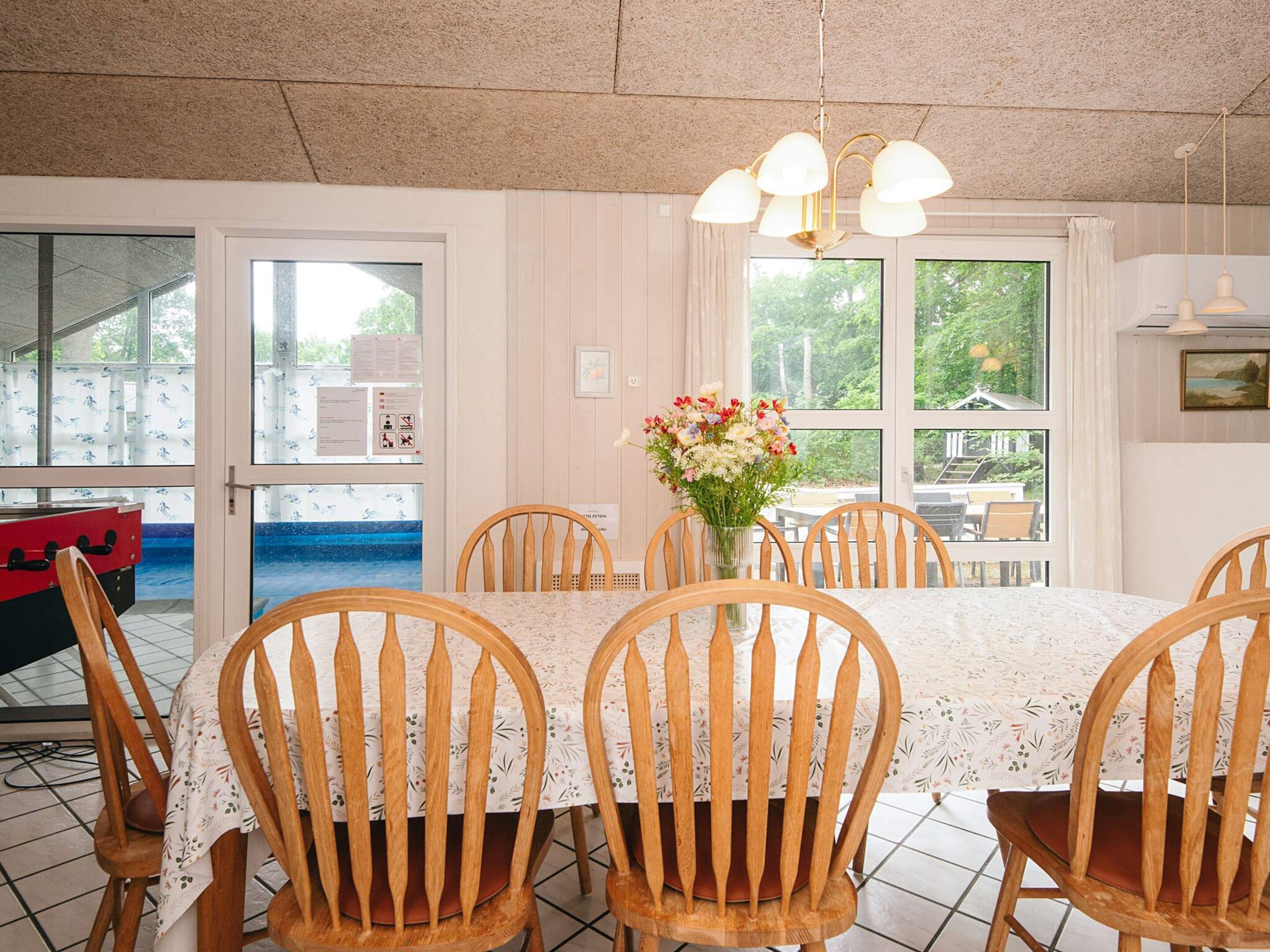 A dining area with a table set for six, surrounded by wooden chairs, a vase of flowers, and a view through large windows.