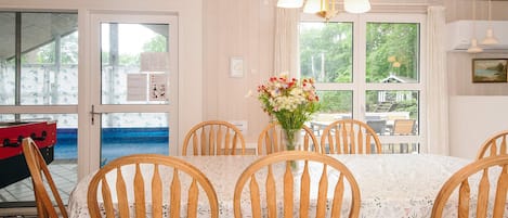 A dining area with a table set for six, surrounded by wooden chairs, a vase of flowers, and a view through large windows.
