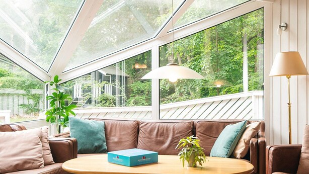 A cozy room with a brown leather sofa, a wooden table, and a large window offering a view of greenery. - 4 Star Holiday Home in Glesborg (Glesborg)