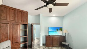 Une chambre avec un parquet en chevrons, un lit, un téléviseur diffusant une scène de plage, un ventilateur de plafond et une armoire.