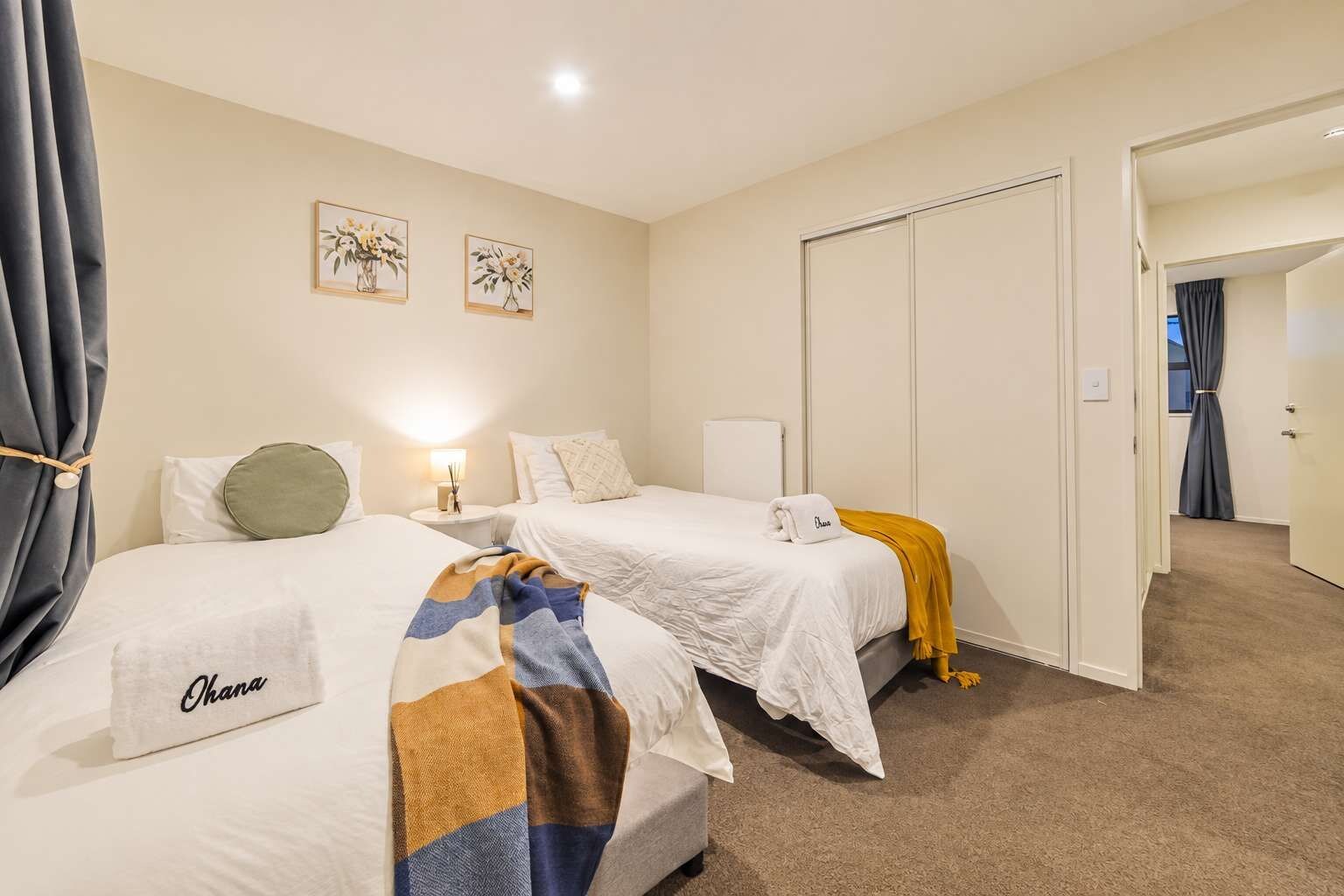 A hotel room with two beds, a bedside table with a lamp, a wall-mounted artwork, and a door leading to another room.