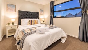A neatly arranged bedroom with a tufted headboard, a large window with curtains, and a bedside table with a lamp.