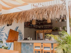A bar area with a thatched roof, wooden chairs, and a shelf with bottles.