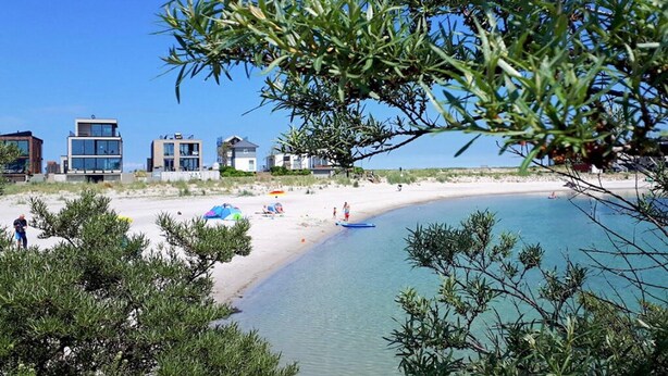 A beach with modern buildings, clear blue water, and sparse vegetation. - Gorgeous apartment in Kappeln with sauna (Kappeln)