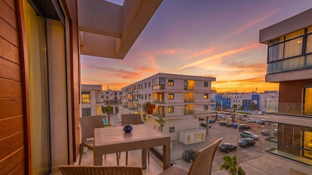 A balcony with wicker furniture, a table with a vase, and a view of modern buildings and a parking area. - Luxury apartment, Agadir bay, pool&fibre (Agadir)