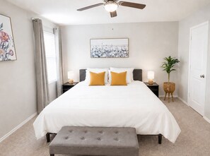 Um quarto com cama, um banquinho, ventilador de teto, mesa de cabeceira, luminária, planta e uma porta.