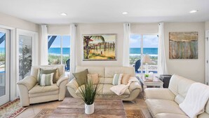Un salon avec un canapé beige, une table basse et une plante en pot. De grandes fenêtres offrent une vue sur la plage.