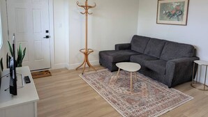 Un salon avec un canapé gris, une petite table blanche, un lampadaire, un tableau accroché au mur et un téléviseur posé sur un meuble.