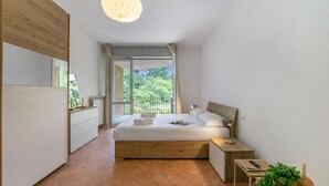 Ein modernes Schlafzimmer mit einem großen Bett, einem Nachttisch, einer Fensterbank und einer Deckenleuchte, mit Blick auf Grünflächen.