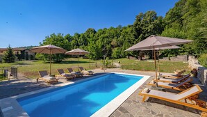 Una piscina amb gandules i ombrelles, envoltada de vegetació i un cel serè.