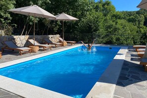 Una piscina con sillas de descanso y sombrillas, rodeada de muros de piedra y árboles.