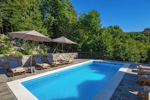 Piscina con sillas de descanso y sombrillas, rodeada de muros de piedra y árboles.