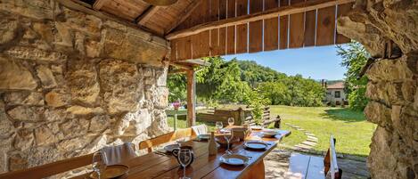 Una mesa de madera al aire libre, dispuesta con platos, vasos y cubiertos, con una pared de piedra y vigas de madera en el techo.