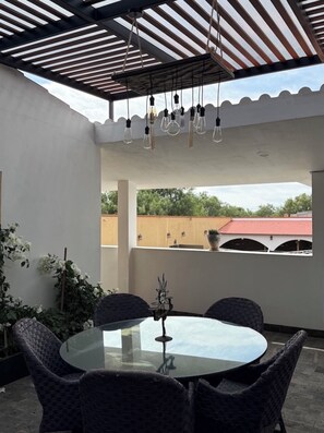 A covered outdoor patio with a round glass table and wicker chairs, a metal candle holder, and a view of a building with a red roof.