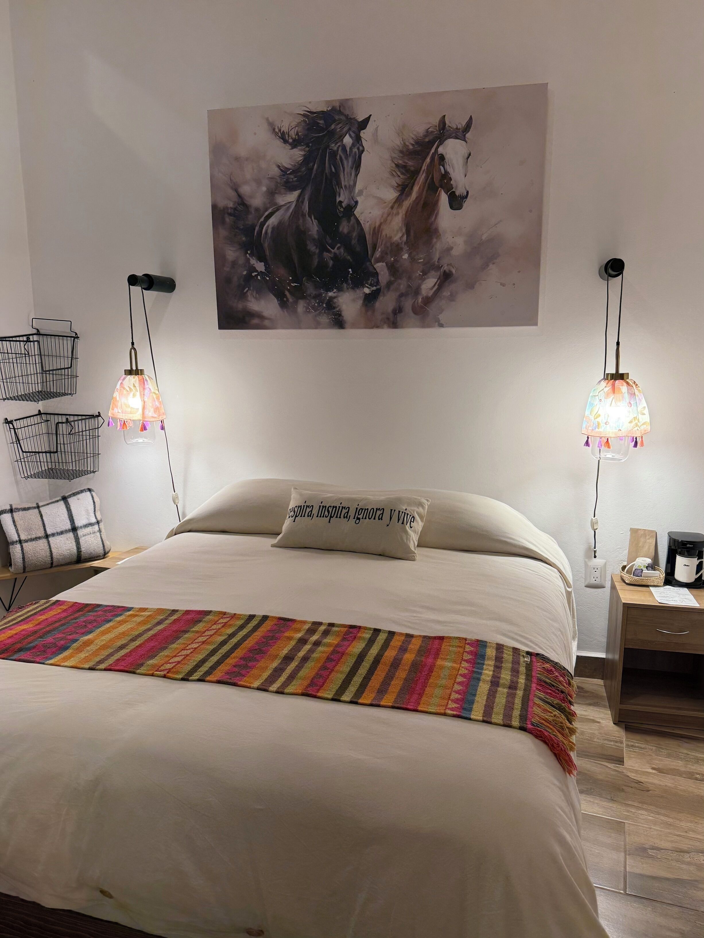 A bedroom with a bed, two wall-mounted lamps, a painting of horses, a pillow with text, and a bedside table with a coffee maker.