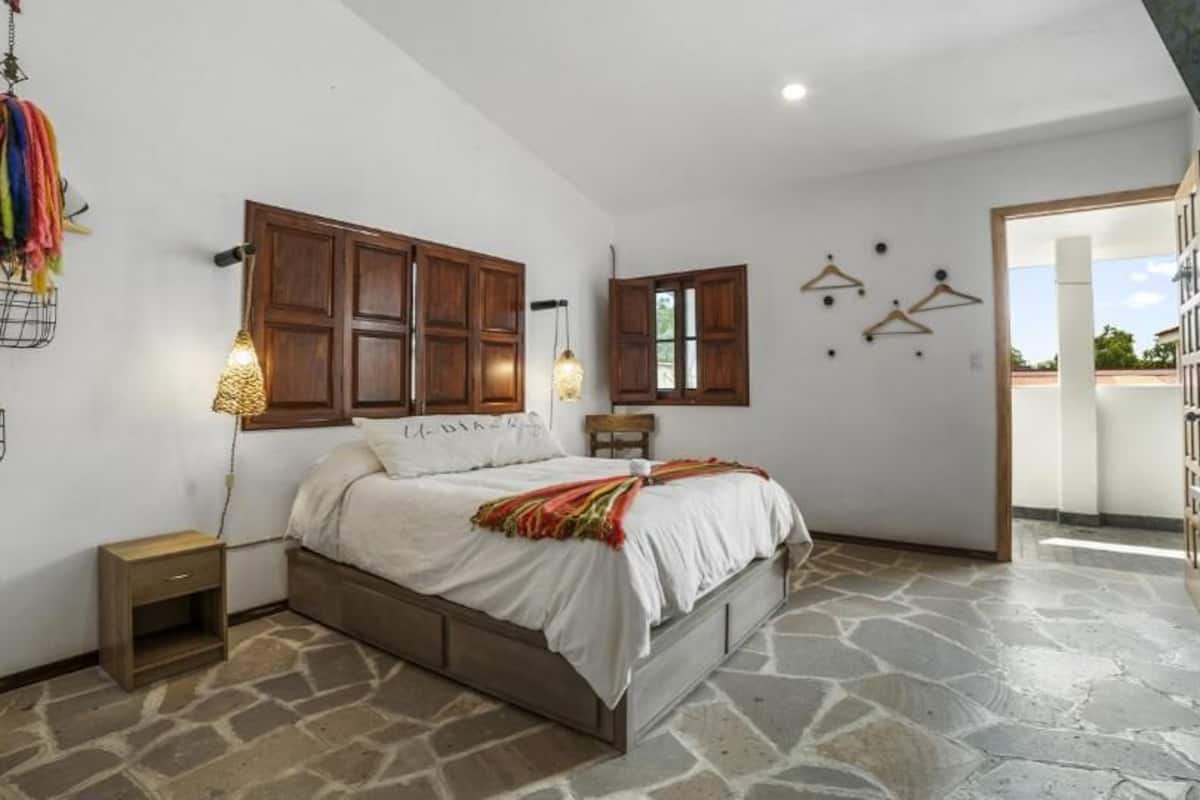 Un dormitorio con suelo de piedra, estructura de cama de madera, mesitas de noche y una ventana con contraventanas de madera.