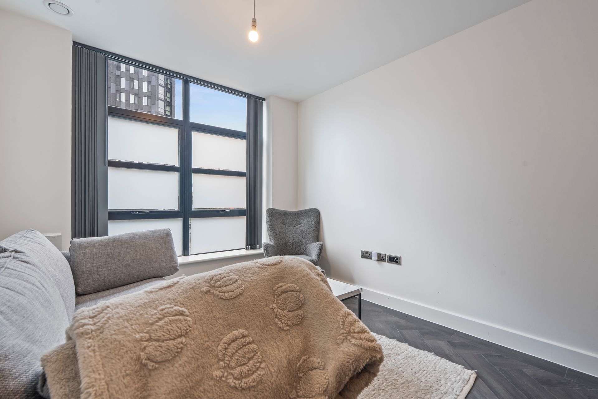 A modern bedroom with a large window, a bed with grey bedding, a fluffy beige throw blanket, and a grey chair.