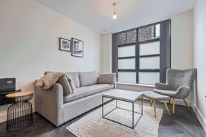 A modern living room with a grey sofa, a coffee table, a grey armchair, a small round table, and a TV. - Stylish 2BDR Loft in Birminghams Jewellery Quarter (Birmingham)