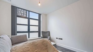 Een moderne slaapkamer met een groot raam, een grijze bank, een beige plaid en een wit vloerkleed.