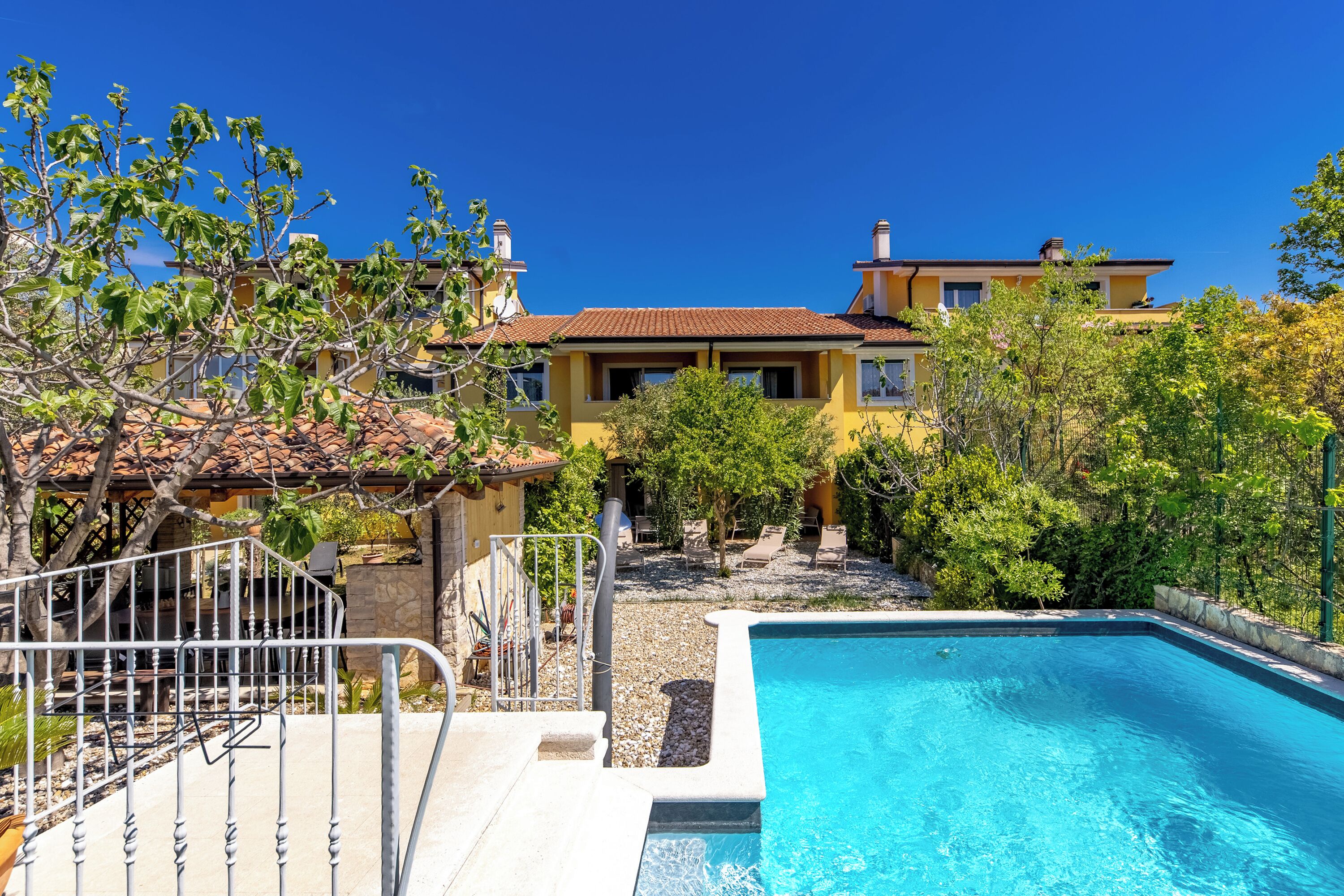 A swimming pool with a stone border, a metal fence, and a yellow building with a red roof in the background.