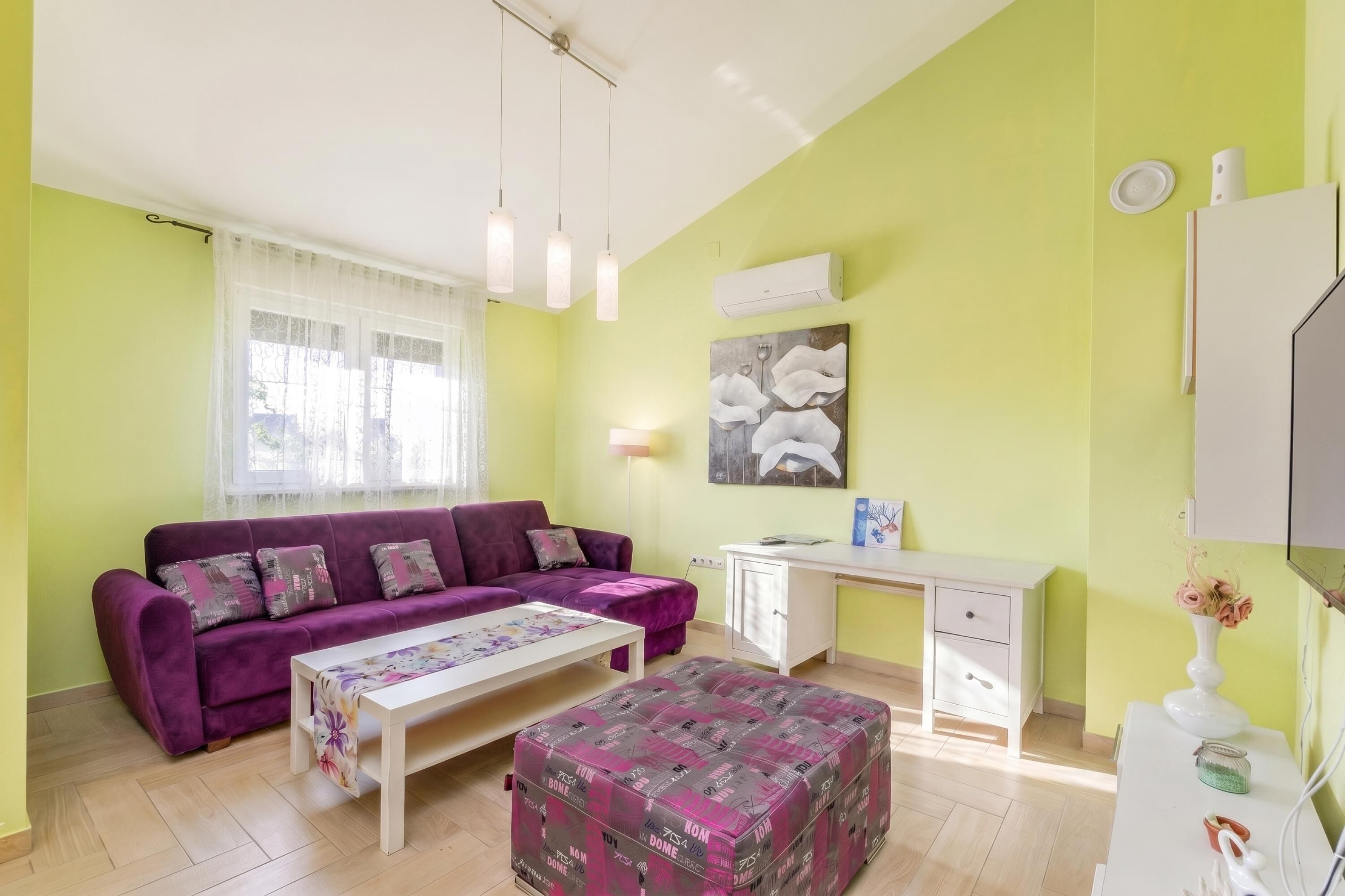 A room with a purple sofa, a white desk, a white cabinet, and a patterned ottoman.