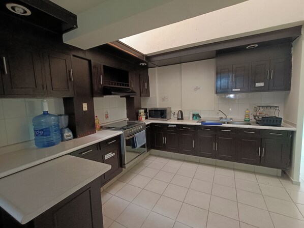 Une cuisine avec des meubles de rangement de couleur sombre, un grand distributeur dâeau, une cuisiniĂšre et un four Ă micro-ondes.